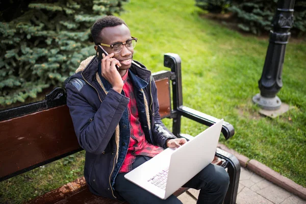Joven empresario que trabaja en el ordenador portátil y hablar de teléfono, sentado en el banco de madera en la calle. Autónomo . — Foto de Stock