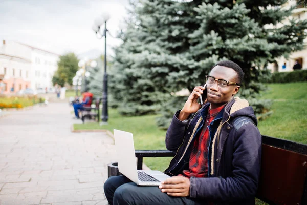 Νέος επιχειρηματίας που εργάζονται για το laptop και να μιλήσουμε τηλέφωνο, κάθεται στο ξύλινο πάγκο στο δρόμο. Ελεύθερος επαγγελματίας. — Φωτογραφία Αρχείου