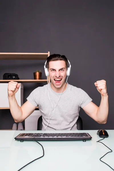Молодой счастливый человек с гарнитурой играет и выигрывает компьютерную игру дома — стоковое фото