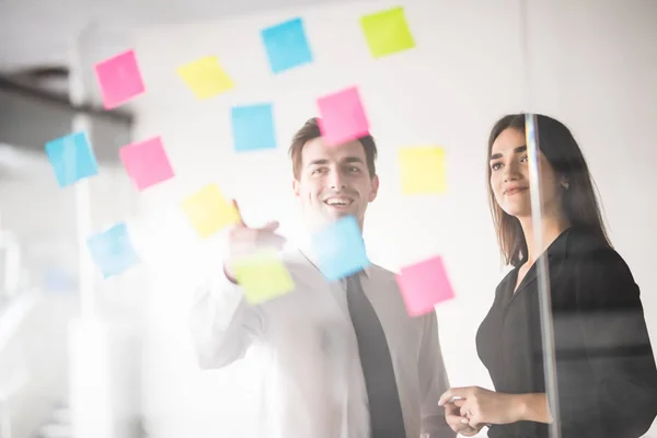 Een man en een vrouw in strikte business kostuums staan op een glazen wand met papieren stickers en kijk goed naar hen. — Stockfoto