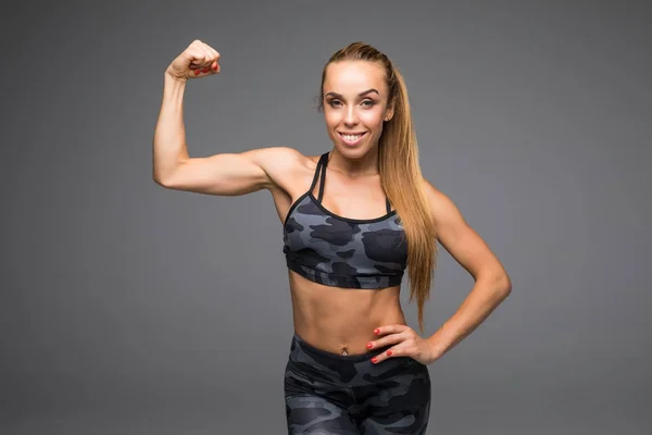 Alegremente sonriente mujer deportiva de raza mixta demostrando bíceps, aislado sobre fondo gris — Foto de Stock