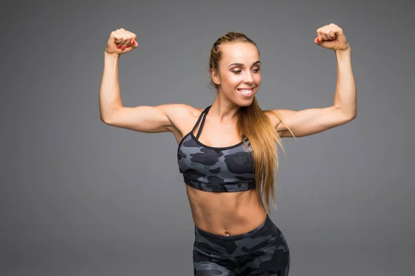 Radośnie uśmiechający się mieszane rasy wykazując biceps sportowy Kobieta, na białym tle na szarym tle — Zdjęcie stockowe