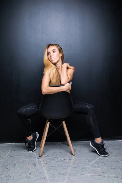 Sexig och vacker ung blond modell med smal kropp poserar i studio på stolen, mörka väggen i bakgrunden — Stockfoto