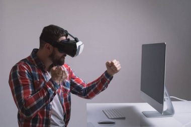 Man in helmet virtual reality plays game at desk. Man uses VR-headset display with headphones for virtual reality game in office in front of monitor. High-tech devices. Augmented reality device creati clipart