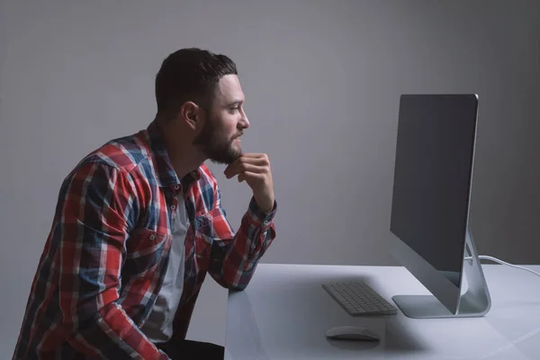 Бородатый кавказский человек сидит за монитором компьютера. Мускулистый красивый парень смотрит на дисплей в рабочее время . — стоковое фото