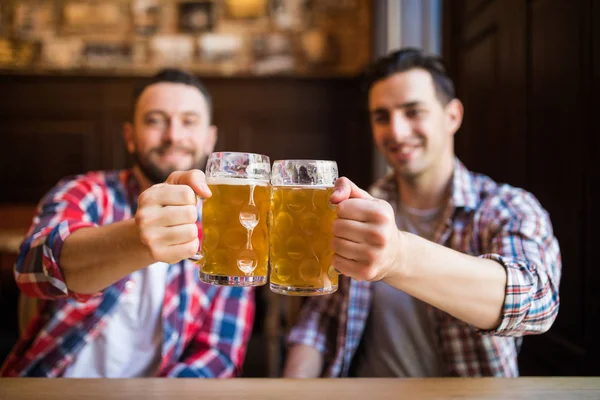 Tomando una cerveza con un amigo. Dos jóvenes alegres brindando con cerveza mientras están sentados juntos en el bar — Foto de Stock