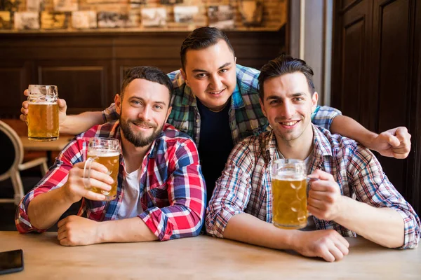 Três jovens em roupas casuais estão sorrindo e batendo copos de cerveja juntos enquanto sentados no pub — Fotografia de Stock