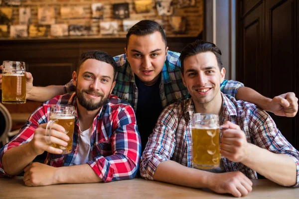 Três jovens em roupas casuais estão sorrindo e batendo copos de cerveja juntos enquanto sentados no pub — Fotografia de Stock