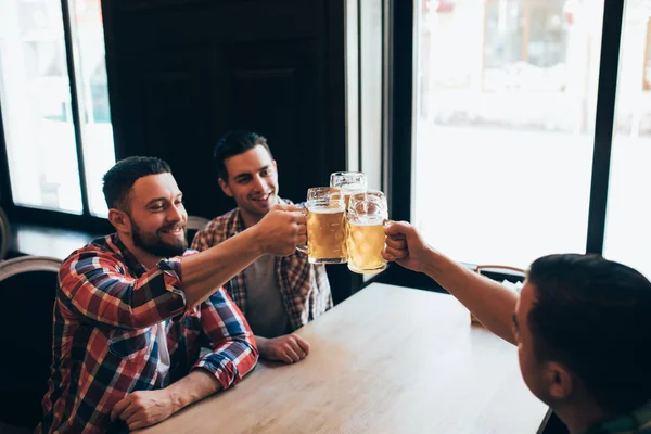 Tres jóvenes hombres felices vestidos con ropa casual sonríen y coquetean vasos de cerveza mientras están sentados en el pub — Foto de Stock