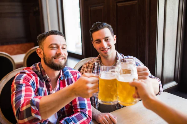 Tre unga lyckliga män i casual kläder leende och klämtande glas öl tillsammans medan du sitter i puben — Stockfoto