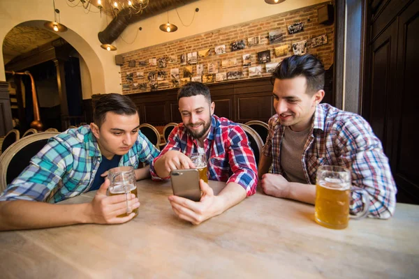 Três jovens em roupas casuais estão conversando, usando um telefone inteligente e bebendo cerveja enquanto estão sentados no balcão do bar no pub. — Fotografia de Stock