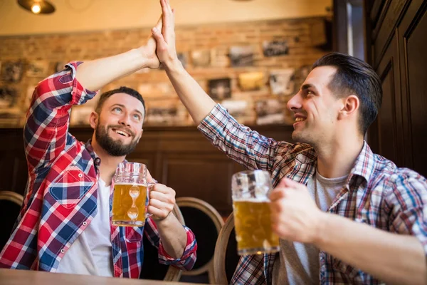 Feliz joven barbudo riendo chocando los cinco con su amigo mientras toman cerveza en el pub juntos — Foto de Stock