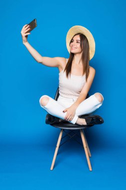 Selfie-portre Studio mavi arka plan üzerinde sandalyede ürpertici şapkalı uzun saçlı güzel kadın yapar.