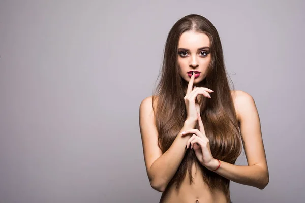 Pretty girl with big eyes and dark eyebrows, with naked shoulders, finger on lips, looking at camera and smiling, a model with light nude make-up, gray studio background — Stock Photo, Image