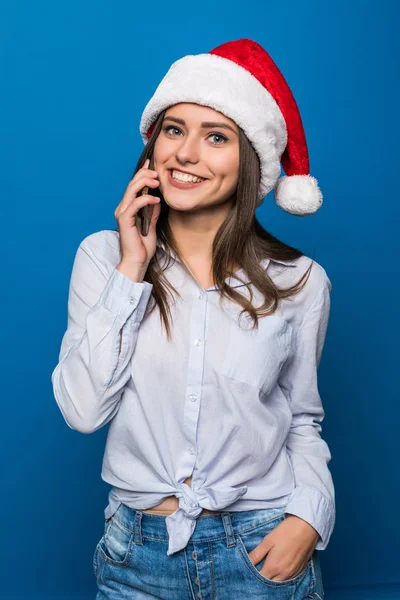 Крупным планом портрет веселой хорошенькой девочки-подростка в шляпе Санты разговаривающей на мобильном телефоне, изолированном на синем фоне — стоковое фото