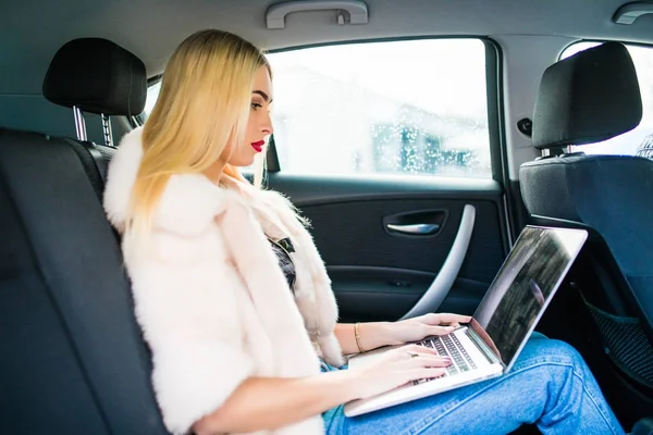 Όμορφη γυναίκα είναι χρησιμοποιώντας ένα φορητό υπολογιστή και χαμογελώντας, ενώ κάθονται στο πίσω κάθισμα στο αυτοκίνητο — Φωτογραφία Αρχείου