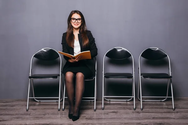 Stanco di aspettare. annoiato giovane donna d'affari in possesso di carta e guardando lontano mentre seduto sulla sedia contro sfondo nero — Foto Stock