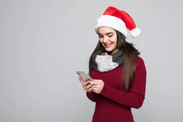 Portret van een brunette jonge vrouw in rode Kerstman met behulp van de mobiele telefoon geïsoleerd op de grijze achtergrond — Stockfoto