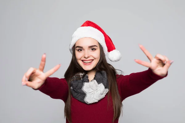Портрет радісної усміхненої дівчини в різдвяному капелюсі, що показує мирний жест і дивиться на камеру ізольовано на сірому фоні — стокове фото