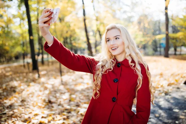 Teenager-Mädchen im Herbst im Park mit einem Selfie auf dem Smartphone. schöne glückliche junge Frau, die ein Foto von sich im Freien an einem Herbsttag macht — Stockfoto