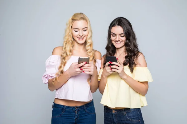 Viciado em Internet. Duas jovens melhores amigas mulheres estão ocupadas em seus telefones celulares em redes sociais, vestindo camisas casuais, isoladas em fundo branco — Fotografia de Stock