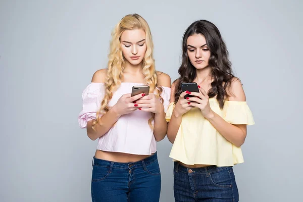 Viciado em Internet. Duas jovens melhores amigas mulheres estão ocupadas em seus telefones celulares em redes sociais, vestindo camisas casuais, isoladas em fundo branco — Fotografia de Stock