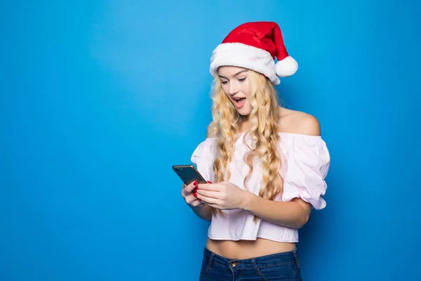 Portret van een lachende mooi meisje gekleed in rode kerstmuts met behulp van mobiele telefoon met verrassing emoties terwijl permanent geïsoleerd op blauwe achtergrond — Stockfoto