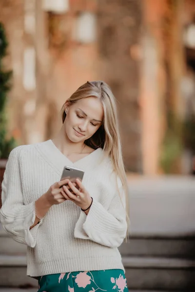 Молодая блондинка пользуется телефоном, на улице. Красивая молодая женщина печатает по телефону в городе в солнечный день . — стоковое фото