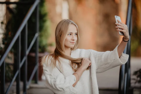 Beautiful casual blonde stylish fashion woman with a phone in her hand make selfie. European buildings and street on a background. Copy space — Stock Photo, Image