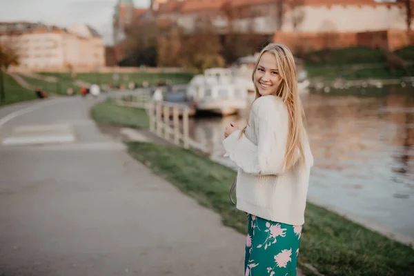 Hermosa mujer joven rubia caminando junto al río al atardecer divertirse sonrisa y jugar en la cámara —  Fotos de Stock