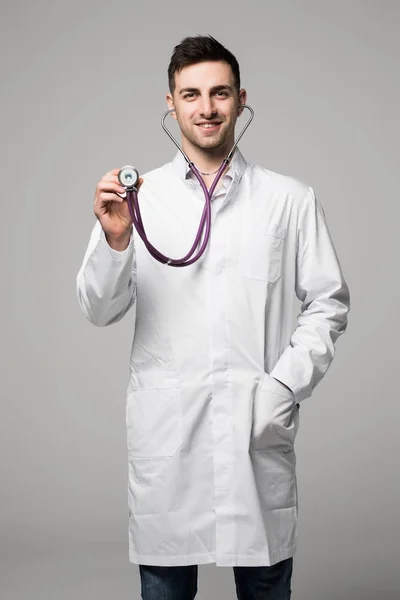 Joven médico guapo sonriente en bata blanca sosteniendo estetoscopio sobre fondo gris —  Fotos de Stock