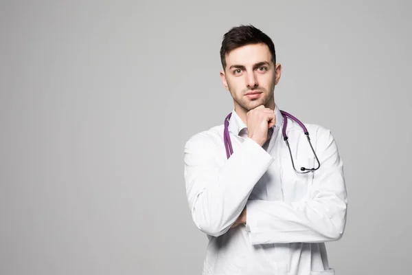 Портрет вдумчивого молодого врача-мужчины со стетоскопом, одетый в униформу, стоящего с рукой на подбородке и смотрящего на пространство для копирования, изолированное на белом фоне — стоковое фото