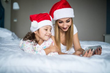 Anne ve kızı santa şapka içinde Noel sabahı yatak ve kullanım telefon portre yatan.