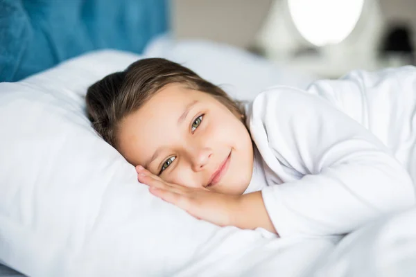 Close-up portrait of laughing little girl lying on bed with hand under pillow, looking at camera and smile. Good morning. — Stock Photo, Image