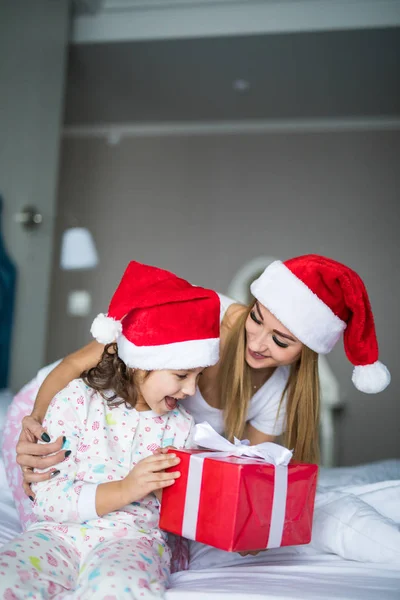 Mutlu bir aile yatakta oturup Noel Baba şapkaları giyiyor. Anne vermek ve Noel hediye ve sahip eğlenceli çocuk Noel zamanında şaşırttı. Kış tatil Noel ve yeni yıl kavramı — Stok fotoğraf