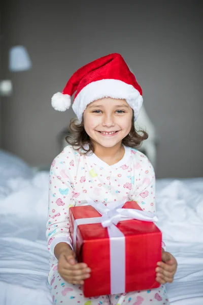 Noel Baba'nın şapkalı kamera gülümseme ile onun hediye kutusu ile beyaz yatakta otururken bakarak çekici çocuk kız — Stok fotoğraf