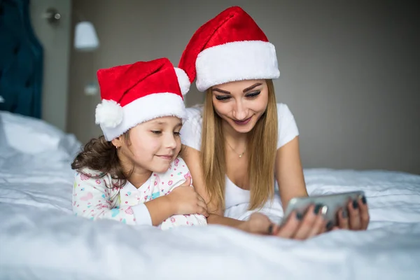Anne ve kızı santa şapka içinde Noel sabahı yatak ve kullanım telefon portre yatan. — Stok fotoğraf