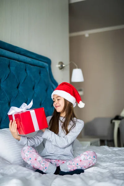 Evde yatakta otururken ona hediye kutusu arıyorsunuz Noel Baba'nın şapkalı büyüleyici küçük kız — Stok fotoğraf