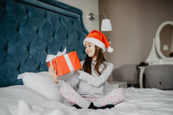 Noel sabahı evde yatakta otururken ona hediye kutusu arıyorsunuz Noel Baba'nın şapkalı büyüleyici küçük kız — Stok fotoğraf