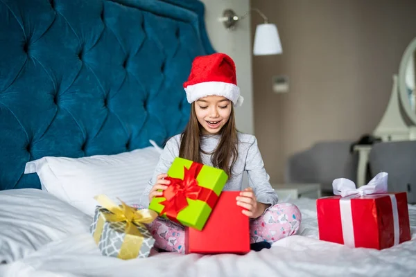 Evde yatakta otururken sürpriz hediye kutusu, seyir açılış mutlu Noel Baba'nın şapkalı güzel küçük kız — Stok fotoğraf
