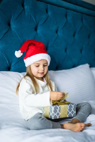 Mutlu Noel Baba'nın şapka hediye kutusu, açılış, küçük kız yatakta otururken gülümseme ile kameraya bakarak çıkıldı — Stok fotoğraf