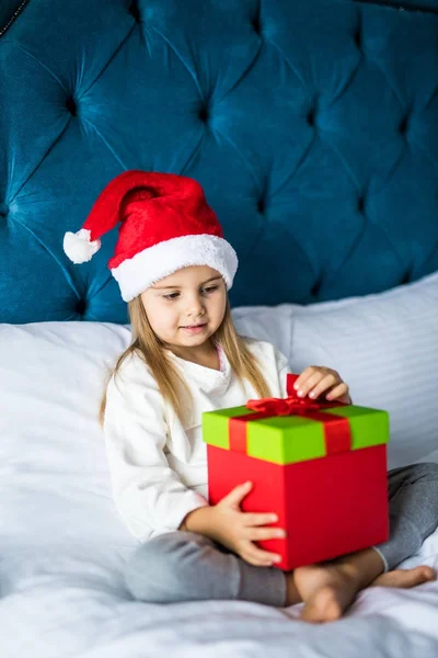 Noel Baba'nın şapka Noel sabahı yatakta otururken ona hediye kutusu dokunmadan, sevimli küçük kız — Stok fotoğraf
