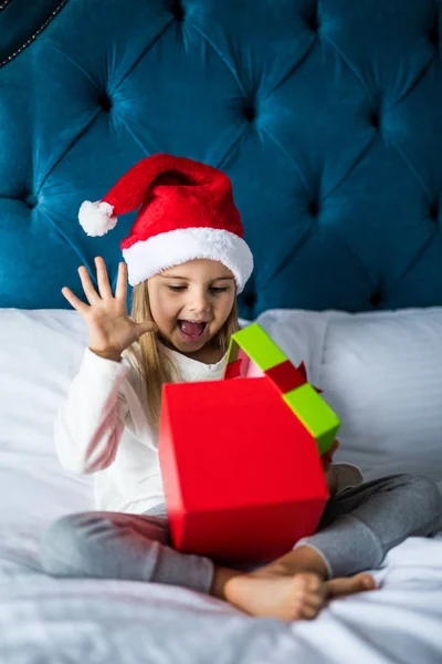 Noel Baba'nın şapkalı sürpriz heyecanlı küçük kız Noel sabahı yatakta çapraz ayaklı otururken mevcut açın. — Stok fotoğraf
