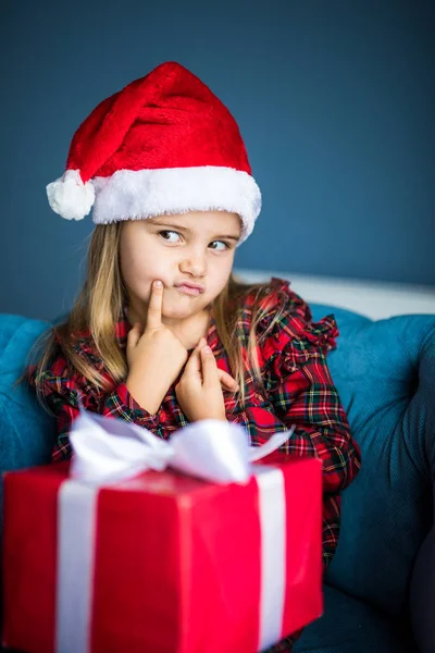 Üzgün kırgın küçük kız evde sandalyede oturup Noel Noel hediye ile elbise ve santa şapka giyiyor — Stok fotoğraf