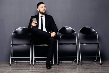 Görüşme için hazır. Yakışıklı adam telefon tutarak resmi kıyafeti içinde ve sandalyeye bekleme odasında otururken kullanabilirsiniz