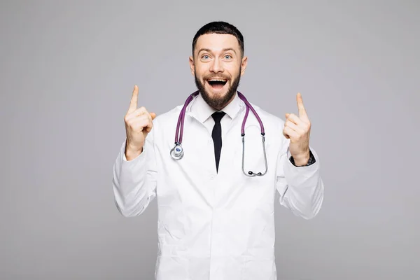 Portréja egy boldog, izgatott férfi orvos öltözött egységes két ujjal mutatva és látszó-on fényképezőgép, elszigetelt fehér háttér felett — Stock Fotó