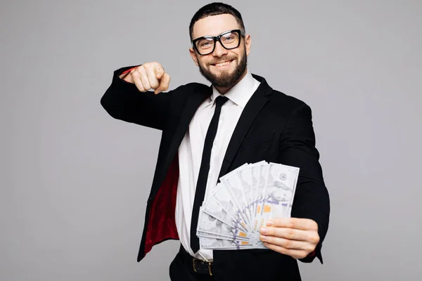 Retrato de um homem sorrindo bem sucedido em terno e óculos apontando o dedo para você com um monte de notas de dinheiro isoladas sobre fundo cinza — Fotografia de Stock