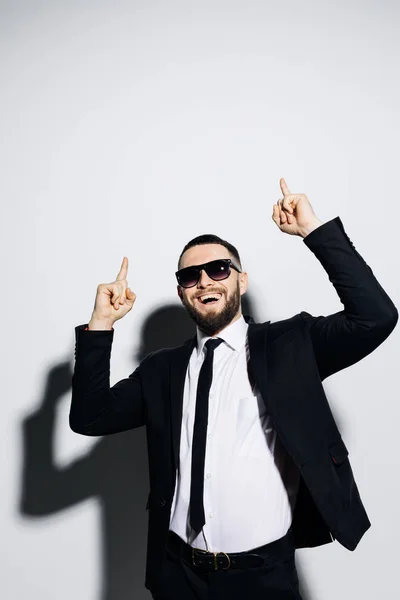 Smiling young business man pointing the finger up while leaning on a grey wall. — Stock Photo, Image