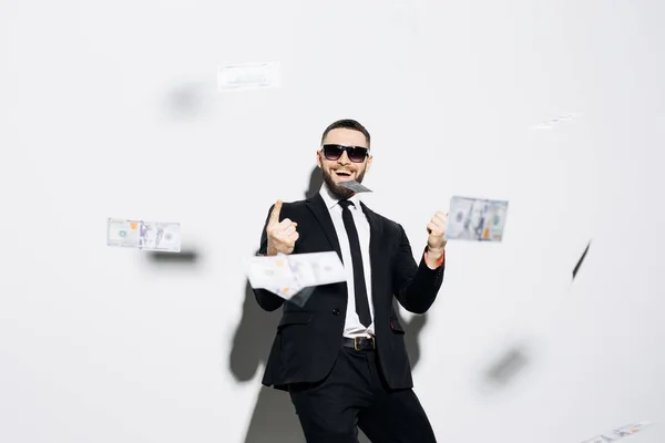 Handsome man in suit and sunglasses pointed up on fluy cash money on white background. Businessman standing in the rain of money. — Stock Photo, Image