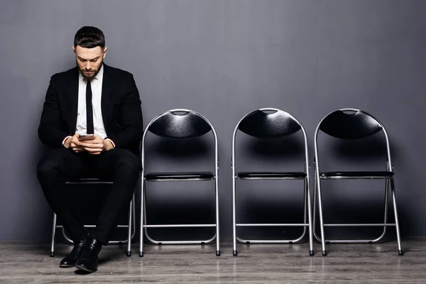 Prêt pour l'interview. Beau homme en tenue de cérémonie tenant le téléphone et l'utiliser tout en étant assis à la chaise dans la salle d'attente — Photo